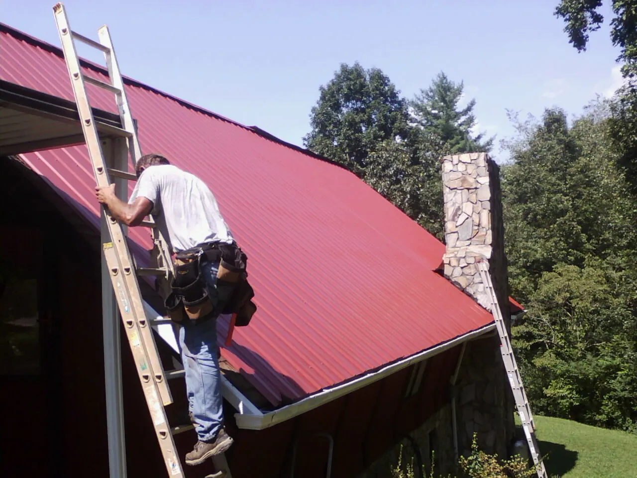 Roofer on a ladder beside a red metal roof and stone chimney — Soffit & Fascia Repair by Roofing Brothers in Nampa